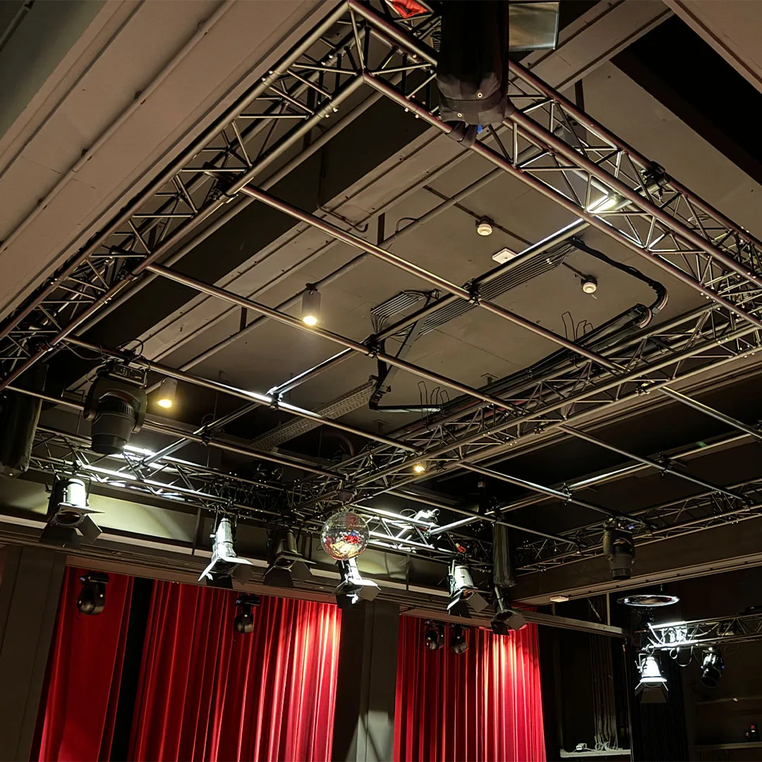 An overhead pipe grid and truss rigged with stage lights and a mirror ball above red drapes